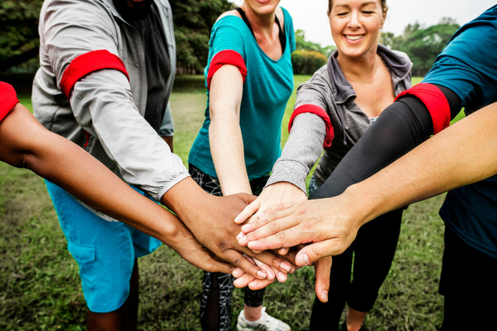 Eine Gruppe Menschen diverser Hautfarben und Geschlechter in unterschiedlichen Outfits stehen im Kreis und halten die Hände in der Mitte zusammen. Sie alle tragen eine rote Armbinde am rechten Arm.