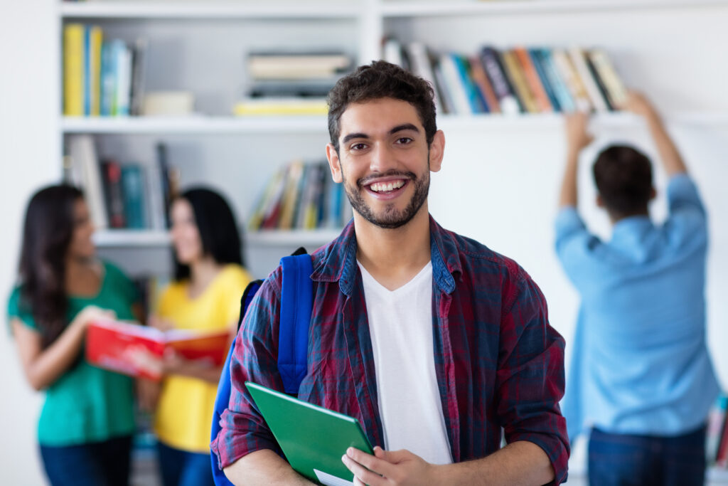 Ein junger Erwachsener mit weißem Shirt und blau-rotem Karohemd steht vor einem unscharfen Bücherregel im Hintergrund. Er befindet sich in einem Raum mit einem Bücherregal und lächelt in die Kamera.